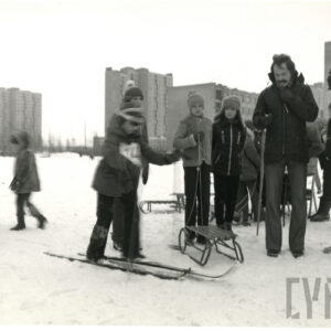 sceneria zimowa, grupa dzieci, część z nich z sankami, z prawej dwóch mężczyzn, w głębi wysokie bloki mieszkalne