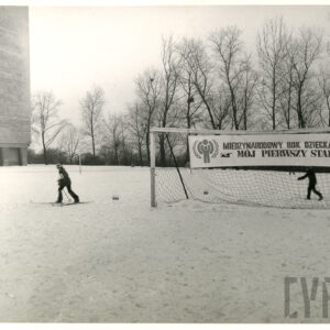sceneria zimowa, z prawej bramka do gry w piłkę nożną oraz dwoje dzieci na nartach, z lewej fragment bloku mieszkalnego