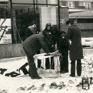 zimowa sceneria, pośrodku grupka młodych ludzi pochylona nad rozłożonym stolikiem, na którym leży tkanina z napisem, w głębi przeszklony pawilon i z prawej fragment bloku
