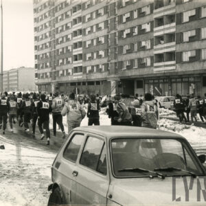 zimowa sceneria, duża grupa ludzi w sportowych strojach i czapkach oraz z numerkami startowymi na plecach biegnie ulicą, na pierwszym planie zaparkowany samochód osobowy, w głębi wysoki blok mieszkalny