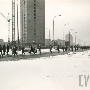 zimowa sceneria, grupa biegaczy w strojach sportowych i z numerami startowymi na piersiach biegnie ulicą, w głębi bloki mieszkalne i dźwig budowlany