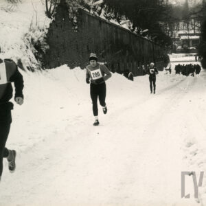 kilku biegaczy w sportowych strojach i czapkach oraz z numerkami na piersiach biegnie ośnieżoną drogą wzdłuż ceglanego muru, w głębi duża grupa biegaczy