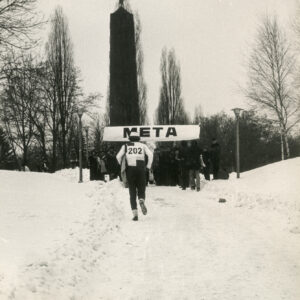 biegacz w sportowym stroju i z numerkiem na plecach dobiega ośnieżoną drogą do szarfy z napisem meta, przy niej grupa ludzi, w głębi wysoki obelisk z gwiazdą na szczycie