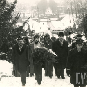 sceneria zimowa, grupa mężczyzn, część trzyma wieńce, wchodzi po ośnieżonych schodach, w głębi ulica i wiadukt kolejowy