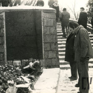 zimowa sceneria, kilku mężczyzn stoi z pochylonymi głowami przed murowaną ścianą z tablicą pamiątkową, pod nią złożone kwiaty, w głębi kilku mężczyzn wchodzi po ośnieżonych, pnących się ku górze kamiennych schodach 