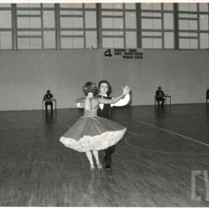 pośrodku sali gimnastycznej tańczy para, pod ścianą, w głębi, przy pojedynczych stolikach siedzą mężczyźni