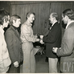 pięciu mężczyzn, pośrodku dwóch mężczyzn zwróconych do siebie twarzami i podający sobie dłonie, jeden z nich trzyma w lewej ręce legitymację