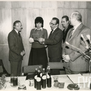 pośrodku stoi kobieta, przy niej kilku mężczyzn, mężczyzna po jej lewej stronie trzyma w ręku płaskie pudełko i pokazuje kobiecie zawartość, na pierwszym planie stół, na nim butelki z napojem, szklanki z napojami i ciastka