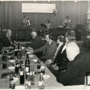 grupa kobiet i mężczyzn siedzi przy stołach, na nich napoje, szklanki, w głębi na scenie zespół muzyczny