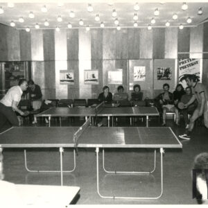 pośrodku sali dwa stoły do tenisa stołowego, przy nich gracze, pod ścianą, w głębi kibice