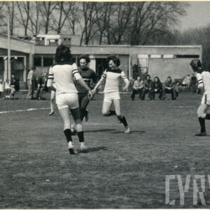 mężczyźni grają na trawiastym boisku w piłkę nożną, przyglądają się im kibice, w głębi parterowy budynek