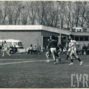 mężczyźni grają na trawiastym boisku w piłkę nożną, przyglądają się im kibice, w głębi parterowy budynek