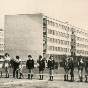 w jednym rzędzie stoją młodzi mężczyźni w strojach sportowych, w głębi pięciokondygnacyjne bloki mieszkalne