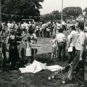 bardzo duża grupa dorosłych z dziećmi częściowo stoi na polanie, częściowo na betonowym boisku