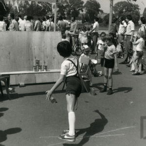 na pierwszym planie chłopiec w krótkich spodenkach celuje do piramidy z puszek, w głębi dzieci w towarzystwie dorosłych