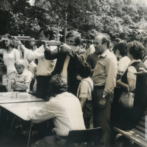 dwóch mężczyzn stoi przy stolikach i celuje z karabinków pneumatycznych, za nimi grupa ludzi