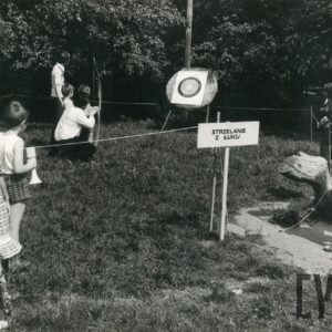 pośrodku polany tarcza strzelecka, z lewej dziecko w towarzystwie mężczyzny szykuje się do strzału z łuku