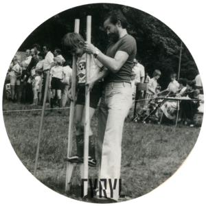 chłopiec idzie na drewnianych szczudłach, pomaga mu mężczyzna, w głębi grupa dzieci w towarzystwie dorosłych