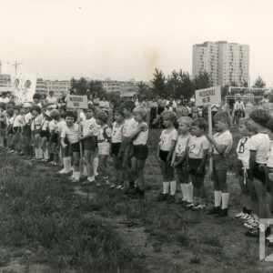 w jednym rzędzie stoją kilkuletnie dzieci w strojach sportowych na trawiastej nawierzchni, w głębi różnej wysokości bloki mieszkalne