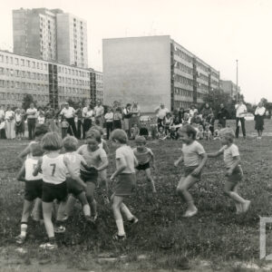 dwie drużyny kilkuletnich dzieci gra w piłkę nożną na trawiastej nawierzchni, przyglądają się grze osoby dorosłe i dzieci, z prawej kobieta w ciemnym sportowym stroju, w krótkich spodenkach przygląda się grze, w głębi kilkukondygnacyjne bloki mieszkalne, za nimi wysoki blok mieszkalny