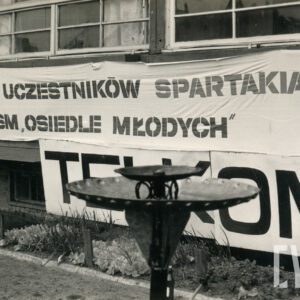 na białej, długiej tkaninie napis „witamy uczestników spartakiady SM Osiedle Młodych”, tkanina zawieszona na budynku, pod oknami parteru