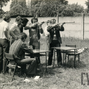 grupa mężczyzn stoi przy dwóch stolikach na trawiastej nawierzchni, dwaj mężczyźni szykują się do strzału z karabinków pneumatycznych, w głębi betonowy płot