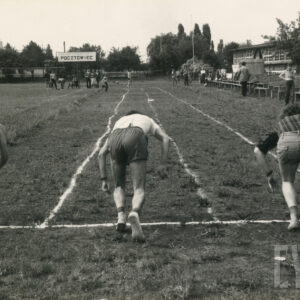 trawiasta bieżnia, na starcie trzech mężczyzn odwróconych tyłem, w sportowych strojach szykuje się do biegu, w głębi grupki kibiców