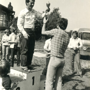 podium medalowe, na nim na zwycięzca zawodów trzyma w ręku puchar, który wcześniej podał mu skierowany w jego stronę mężczyzna, wokół grupa ludzi