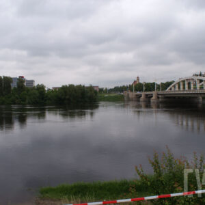 most łączący dwa brzegi, pod nim rzeka o wyraźnie podniesionym stanie; w oddali zarysy budynków