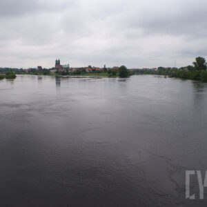rzeka o podniesionym stanie, w oddali wyspa z zarysami budynków; po obu stronach rzeki drzewa oraz zabudowania