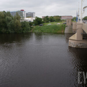 most łączący dwa brzegi, pod nim rzeka o wyraźnie podniesionym stanie; w oddali zarysy budynków