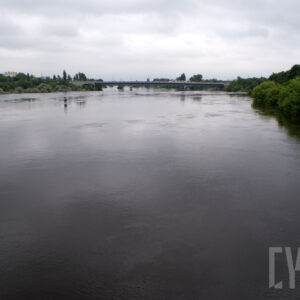 na pierwszym planie rzeka; po lewej gęsto rosnące drzewa oraz zabudowania; po prawej gęsto rosnące drzewa; w tle most oraz zarys zabudowań