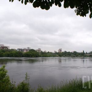 na pierwszym planie rzeka; po prawej most oraz zarys drzew; na drugim planie gęsto rosnące drzewa oraz zabudowa