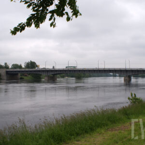 na pierwszym planie rzeka oraz fragment przybrzeżnej łąki; po lewej drzewa oraz zarys zabudowy; po prawej zarys łąki oraz drzew; w tle most oraz zarys drzew
