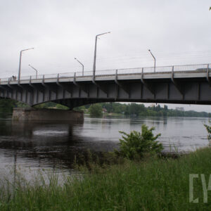 na pierwszym planie rzeka oraz most; po prawej zarys łąki oraz drzew; w tle gęsto rosnące drzewa oraz zarys zabudowań