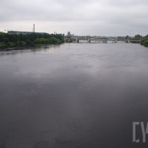 na pierwszym planie rzeka; na obu brzegach drzewa; dalej most oraz zarys zabudowań