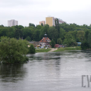 na pierwszym planie rzeka; na drugim planie gęsto rosnące drzewa, zarys dwóch budynków, 5 osób oraz dwa samochody policyjne; w tle gęsto rosnące drzewa oraz zarys zabudowy 