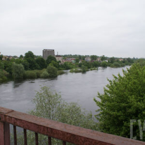 na pierwszym planie rzeka oraz fragment barierki mostu; po prawej stronie gęsto rosnące drzewa; w tle gęsto rosnące drzewa oraz zabudowania