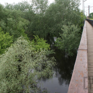 na pierwszym fragment drewnianej kładki dla pieszych wzdłuż stalowej konstrukcji mostu, pod nim rzeka oraz wystające ponad poziom wody korony drzew