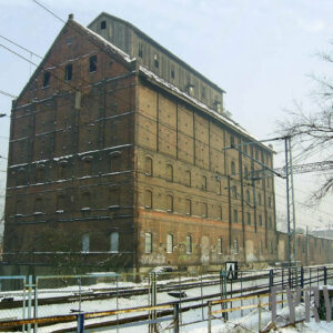 kilkukondygnacyjny, przemysłowy budynek ceglano-drewniany ze spadzistym dachem i drewnianą nadbudówką na dachu, większość otworów okiennych zamurowana, przed nim tory kolejowe i sieć trakcyjna