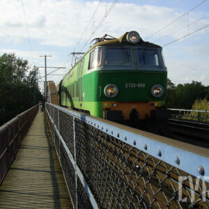 żelazny most kratownicowy z torami kolejowymi, siecią trakcyjną i kładką dla pieszych, na moście ciężka lokomotywa elektryczna, z lewej i prawej strony drzewa