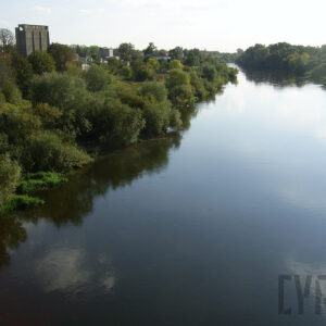 na pierwszym rzeka z dzikim porośniętym drzewami i trawą brzegiem, w głębi z lewej pozostałości betonowego, wysokiego budynku, z prawej od niego przebijające się znad drzew niskie budynki mieszkalne