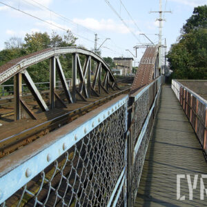 żelazny most kratownicowy z torami kolejowymi, siecią trakcyjną i kładką dla pieszych, z lewej i prawej strony drzewa, w głębi kilkukondygnacyjny budynek ze spadzistym dachem i niższą dobudówką 