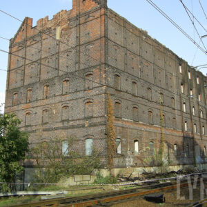 kilkukondygnacyjny, przemysłowy budynek ceglano-drewniany z zamurowanym otworami okiennym i z całkowicie spalonym dachem, przed nim żelazny most kratownicowy z torami kolejowymi i siecią trakcyjną