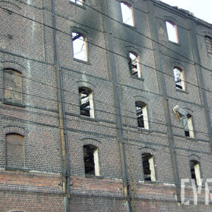 ściana kilkukondygnacyjnego, przemysłowego budynku ceglanego z widocznymi śladami po pożarze, większość pustych otworów okiennych, przez które widać wypaloną, drewnianą konstrukcję wnętrza