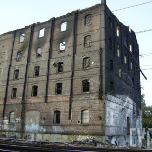 kilkukondygnacyjny, przemysłowy budynek ceglany z widocznymi śladami po pożarze, większość pustych otworów okiennych, przez które widać wypaloną, drewnianą konstrukcję wnętrza