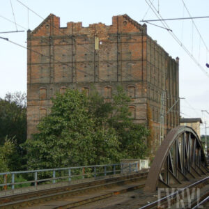 kilkukondygnacyjny, przemysłowy budynek ceglano-drewniany z zamurowanym otworami okiennym i z całkowicie spalonym dachem, przed nim żelazny most kratownicowy z torami kolejowymi i siecią trakcyjną