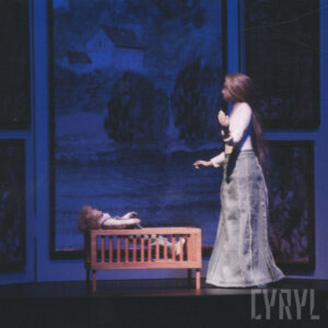 lalka kobiety, przy niej łóżeczko, w nim lalka chłopca, za nimi fragment malowanej scenografii