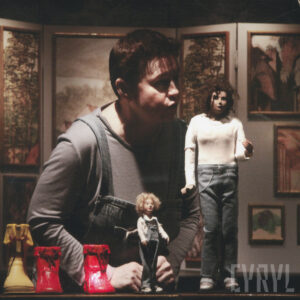 lalki mężczyzny i chłopca, przy nich z lewej trzy buciki, za nimi aktorka animująca lalkami, w głębi powieszone obrazy jako element scenografii