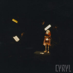 pośrodku, w prawie całkowitych ciemnościach na czarnym podwyższeniu stoi oświetlona lalka, w głębi widoczne płaskie podświetlone elementy, które bardzo lekko oświetlają twarze kilku aktorów